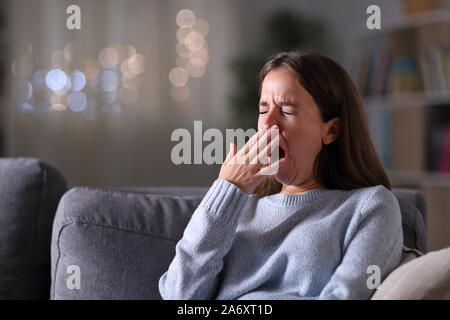 Tired woman yawning couvrant la bouche avec une main assis sur un canapé dans la nuit à la maison