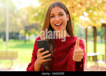 La mode femme attrayante à l'aide de smart phone montre thumb up en plein air, sur l'automne. Banque D'Images