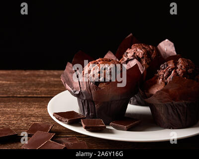 Still Life with brown muffins au cacao dans un emballage de papier cuisson, les morceaux de chocolat sur une assiette de porcelaine blanche et des dessus de table. Black Banque D'Images