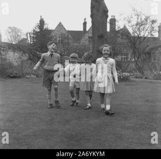 Années 1950, historique, quatre jeunes enfants heureux bien habillés tenant la main dehors sur la pelouse d'un jardin clos d'un manoir, Angleterre, Royaume-Uni, ayant assisté à un service du dimanche à l'église du village. Banque D'Images