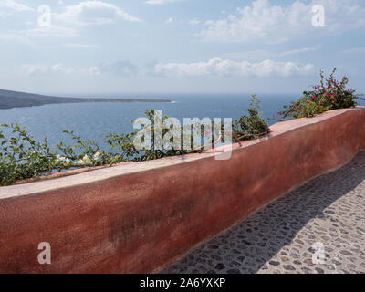 Une longue diagonale rouge ocre et mur en pierre et masse pavés de galets d'arrière-plan des montagnes avec une mer calme sous ciel bleu avec cloud Oia Santorini Banque D'Images