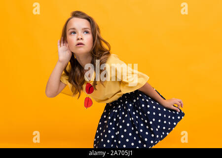 Petite fille eavesdrops sur une conversation, un enfant est titulaire d'un palm par l'oreille contre l'arrière-plan d'un mur orange Banque D'Images