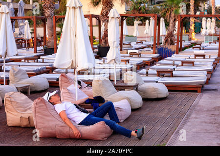 Voyageur fatigué se repose sur un oreiller de plage parmi les recueillies parasols de plage avec des chaises longues sur une plage vide sous les palmiers. Banque D'Images