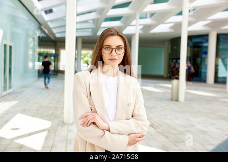 Portrait de la belle young businesswoman looking at camera et souriant tout en se tenant à l'extérieur, près de l'immeuble de bureaux modernes. Banque D'Images