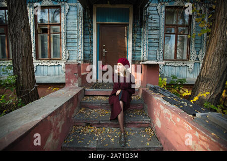 Femme dans un manteau de bourgogne et beret attend assis sur le perron d'une vieille maison en bois. Banque D'Images