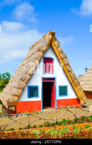 Maison traditionnelle dans la région de Santana, l'île de Madère, au Portugal. Les petites maisons en bois, triangulaire, avec toit de chaume et façade colorée. Partie de patrimoine portugais. Jardin de fleurs en premier plan. Journée ensoleillée. Banque D'Images