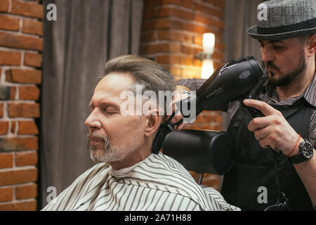 Grandpa obtient une coupe chez le coiffeur dans un salon de coiffure. Coupe de cheveux à la mode d'un vieil homme, un pensionné dans le barber shop. Soins des cheveux et de la barbe. La Russie, S Banque D'Images