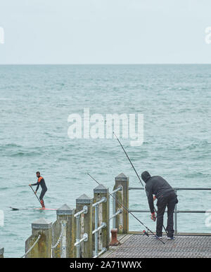 Un pêcheur ajuste sa verge sur le pont de pêche de la jetée de Bournemouth, Angleterre, comme un homme sur un paddle board laissez-passer. Banque D'Images