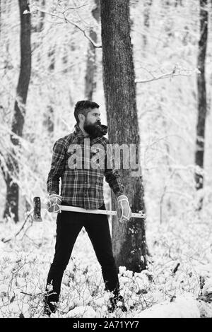Homme avec gants chauds et veste à carreaux en forêt. Barbe et moustache avec macho est titulaire d'ax. Guy avec la lettre, avec des arbres couverts de neige sur l'arrière-plan. Hipster woodsman concept. Banque D'Images