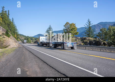 Gros camion bleu classique American semi truck transport de deux semi-remorques à fond plat vide passe à l'entrepôt pour charger d'exécution sur la route le long de la Colombie-Britannique Banque D'Images