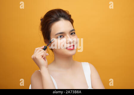 Closeup portrait of attractive young woman putting mascara certains sur ses cils avec pinceau maquillage sur fond jaune Banque D'Images