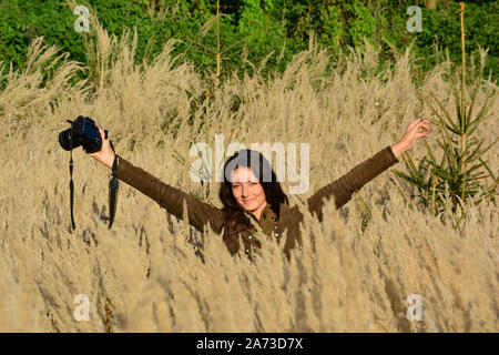 Jeune fille brune aux cheveux longs, photographe de la nature dans les hautes herbes de couleur d'or, au milieu de l'automne, à la bonne humeur après travail bien fait Banque D'Images