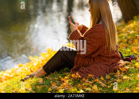 Jeune femme textos un message sur son téléphone portable pendant qu'elle se détend dans un parc assis sur le gazon entouré de feuilles aux couleurs automnales Banque D'Images