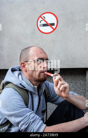 La fumée de cigarette électronique man en vertu de l'inscription ne pas fumer au city street. Violation des règles au concept de lieu public. Iqos en main système Banque D'Images