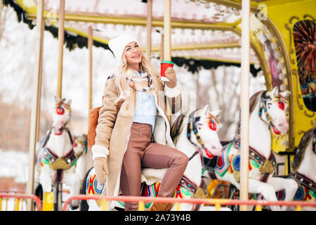 Une belle jeune femme blonde en peau de mouton d'hiver élégant manteau, gants, Chapeau tricoté, avec sac à dos, équitation manège, tenant un café chaud Banque D'Images