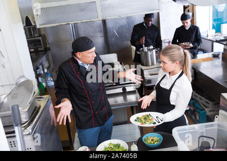 Jeune serveuse debout avec plat cuisiné dans une cuisine de restaurant à parler avec perplexe male chef Banque D'Images