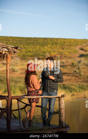 Couple buvant un verre de vin sur un ponton en bois avec lac derrière elle au coeur d'un grand vignoble. Banque D'Images