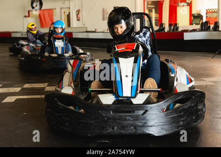 Jeune femme au casque et d'autres personnes qui conduisent des voitures en go-kart indoor sport club Banque D'Images
