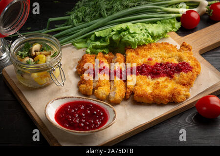 Escalope de poulet sur une planche avec des fines herbes et sauce aux airelles dans une tasse, servi avec pommes de terre sautées aux champignons Banque D'Images
