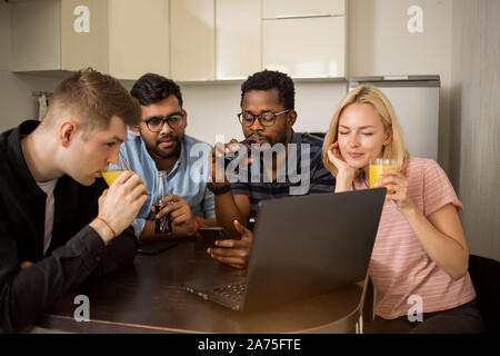 Groupe d'étudiants de diverses assis à table, boire du jus, à l'aide de téléphone mobile et ordinateur portable, regardant la vidéo ensemble. Quatre amis s'amuser dans la cuisine Banque D'Images