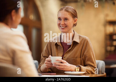 Jeune femme assise à la table avec une tasse de café et souriant à son ami alors qu'ils sont in cafe Banque D'Images