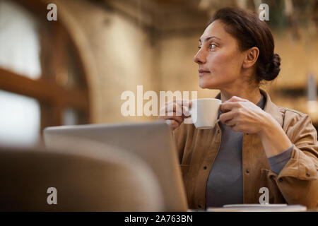 Mature Woman holding tasse de café et à l'écart avec la vue tout en restant assis et pensif working on laptop Banque D'Images