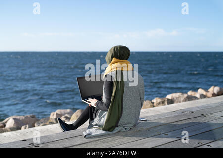 Jeune femme portant le foulard, assis près d'ocean using laptop Banque D'Images