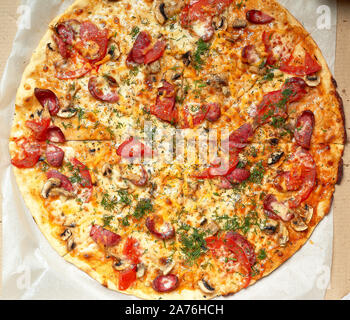 Ronde au four pizza avec saucisses fumées, champignons, tomates, fromage et de l'aneth dans une boîte en carton sur une table en bois, de l'alimentation est coupée en portions, t Banque D'Images