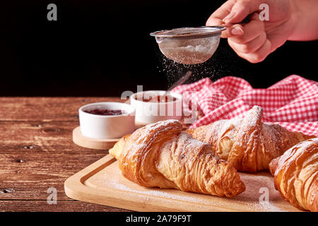 La vie encore sur de vieux tapis en bois avec trois croissants et de la confiture de fruits confits à la pétanque. Main tenant un tamis de sucre sur un fond noir. Banque D'Images