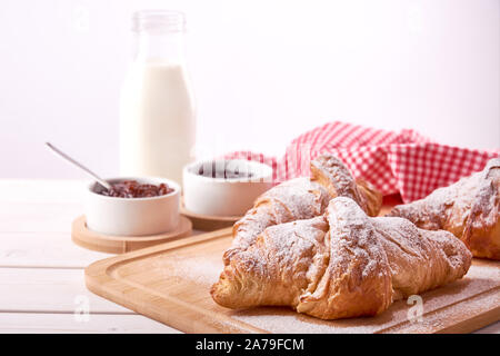 La vie encore sur la vieille planche de bois table avec trois croissants et de la confiture de fruits doux. Verre de lait sur fond blanc. Banque D'Images