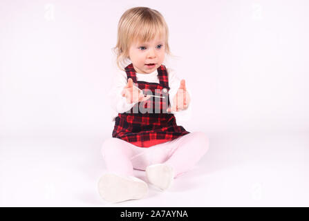 Petite blonde bébé fille avec de grands yeux bleus en robe rouge assise sur le sol, fond blanc. Portrait isolé Banque D'Images