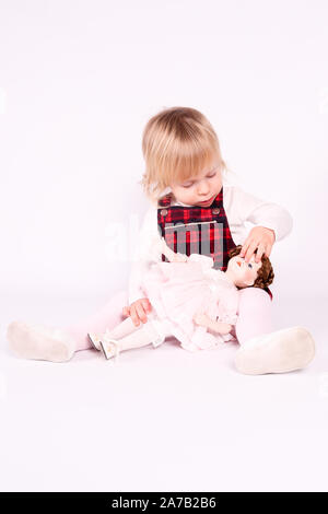 Petite blonde bébé fille avec de grands yeux bleus en robe rouge jouer avec l'ancienne poupée. Assis sur le plancher, fond blanc. Portrait isolé Banque D'Images