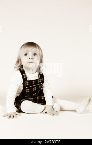 Petite blonde bébé fille avec de grands yeux bleus en robe rouge assise sur le sol, fond blanc. Portrait isolé Banque D'Images