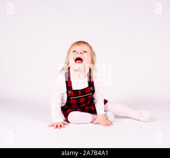 Petite blonde bébé fille avec de grands yeux bleus en robe rouge assise sur le sol, fond blanc. Portrait isolé Banque D'Images