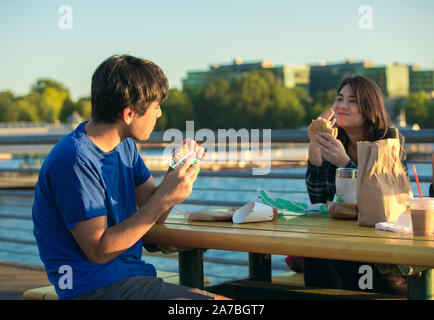 Deux jeunes asiatiques biracial, caucasien adultes assis à l'extérieur par le lac en zone urbaine de manger des hamburgers et de parler ensemble à une table de pique-nique près de Sunset Banque D'Images