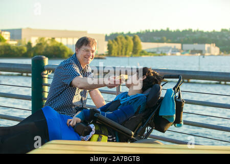 Père de race blanche au début des années 50 en mobilité d'alimentation fils de treize ans dans un hamburger en fauteuil roulant extérieur dès les beaux jours par lake Park en zone urbaine Banque D'Images