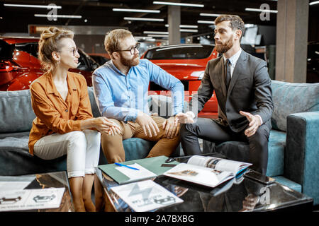 Jeune couple avec directeur des ventes sur le canapé confortable à l'agence de voiture. Jeune famille d'acheter la voiture électrique sur certaines choses formelles avec le vendeur dans la salle d'exposition Banque D'Images