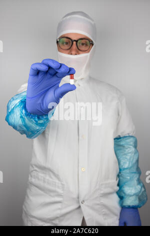 Jeune femme avec des lunettes portant des vêtements de protection stérile blanc et bleu gants tenant une pilule rouge et blanc en face d'elle Banque D'Images