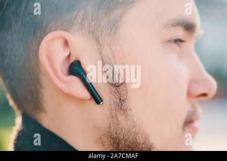 Bichkek, Kirghizistan - le 10 octobre 2019 : Jeune homme dans un manteau et sans fil casque personnalisé apple noir airpods se trouve dans le parc sur un banc Banque D'Images