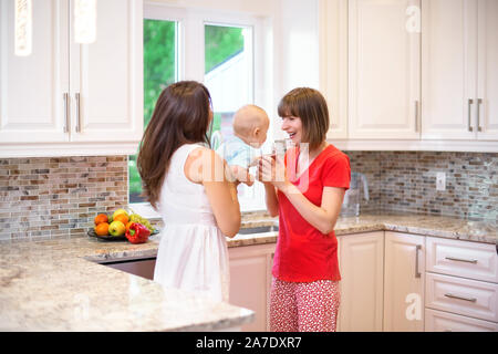 La notion de maternité, nanny, la petite enfance et l'enfance. Une balle dans la cuisine. Deux femmes et un enfant dans leurs bras, l'enfant reçoit l'eau, une Banque D'Images