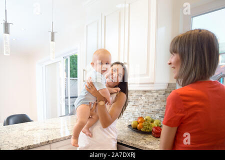 La notion de maternité, nanny, la petite enfance et l'enfance. Une balle dans la cuisine. Deux femmes et un bébé dans leurs bras, à la recherche dans l'appareil photo et s Banque D'Images