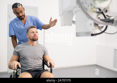 Jeune médecin en uniforme montrant homme malade de nouveau matériel médical fauteuil roulant Banque D'Images