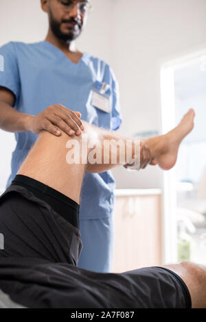 Médecin spécialiste contemporain en uniforme tenant la jambe malade de son patient Banque D'Images