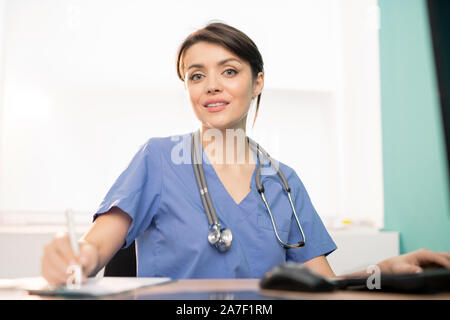 Pretty young smiling doctor en uniforme bleu de travail par la prise de notes Banque D'Images