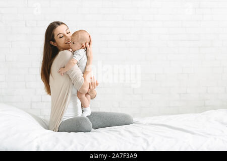 Young woman embracing adorable baby sitting on bed at home Banque D'Images