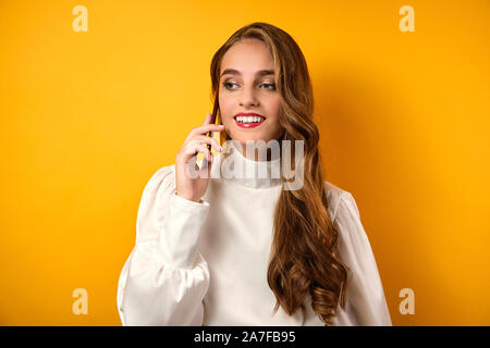 Une belle fille avec des boucles et le rouge à lèvres rouge dans un chemisier blanc se dresse sur un fond jaune et parle au téléphone. Banque D'Images