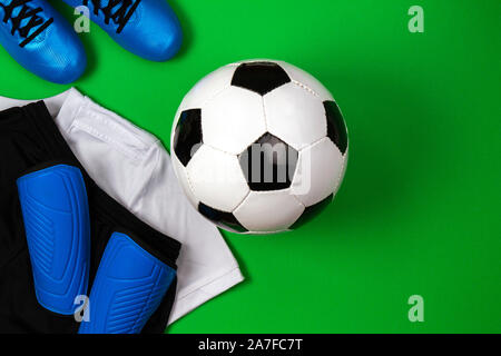 Ballon de soccer, bottes bleues, taquets, t-shirt blanc et short noir sur fond vert. Mise à plat, vue du dessus Banque D'Images