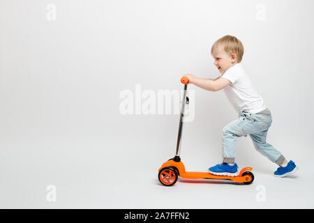 Heureux tout-petit enfant dans une orange en jeans scooter sur un fond blanc dans le studio. La publicité pour les jouets pour enfants avec l'espace pour le texte. Banque D'Images