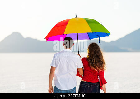 Dos de jeune couple romantique parapluie arc-en-ciel colorés pour l'ombre sur la Bonneville Salt Flats blanc près de Salt Lake City, Utah et sur la montagne Banque D'Images