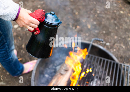 Femme tenant un plateau/bouilloire de l'eau mise sur le gril à feu au camping avec flamme rouge feu brûler en Soir ou matin avec mitten Banque D'Images